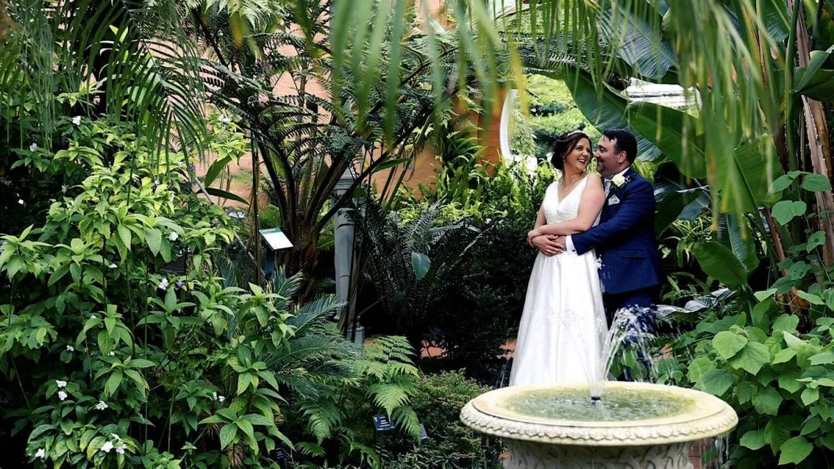Bride in a white dress and groom in a navy suit embracing among tropical plants beside a stone fountain