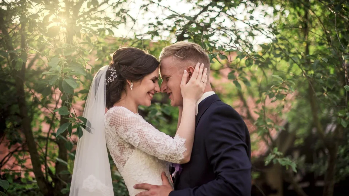 Bride and groom embracing under sunlight with trees in the background.