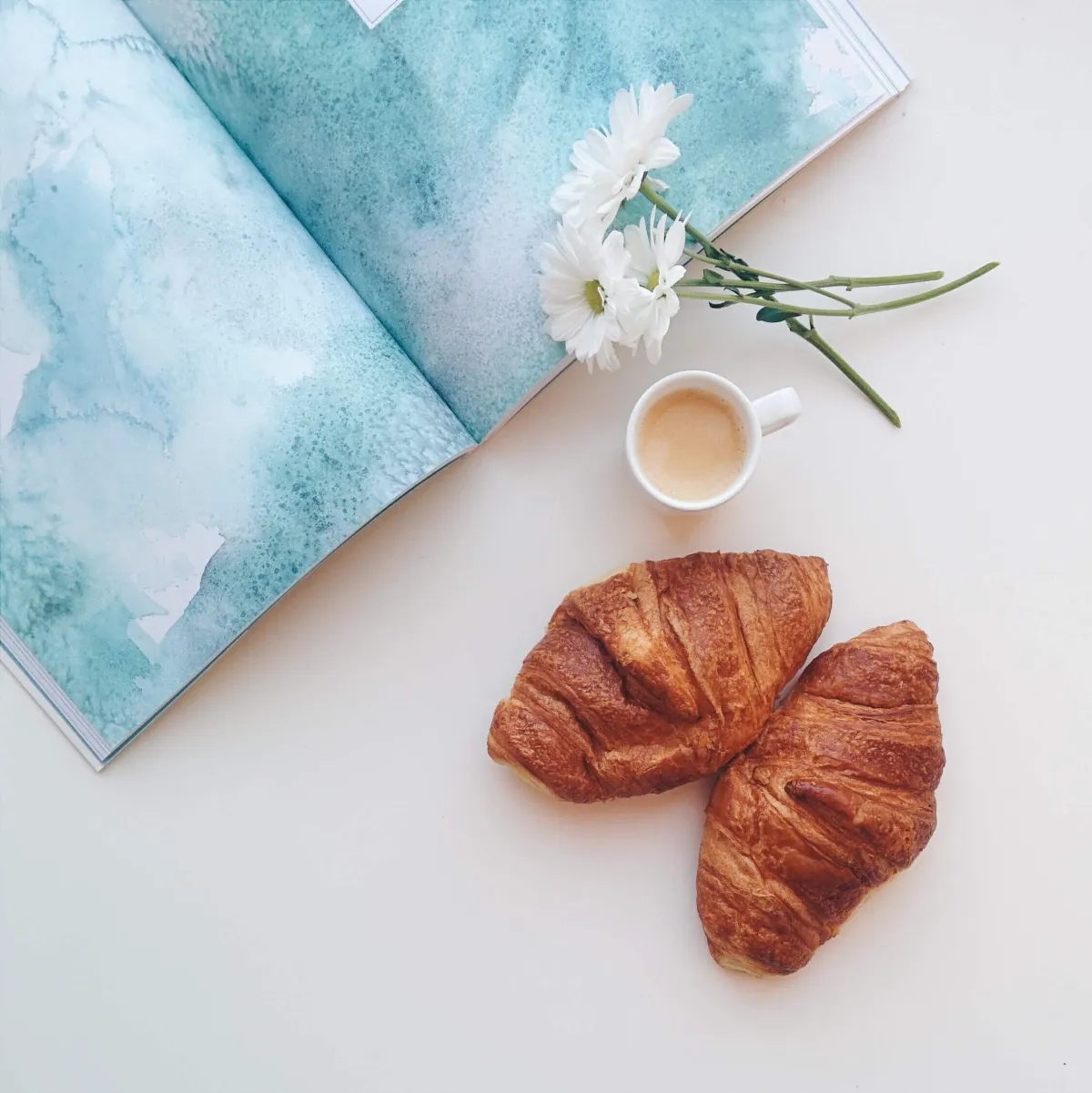Two croissants, a cup of coffee and white daisies beside an open magazine