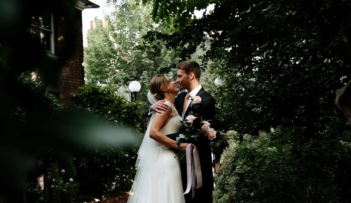 Bride and groom embracing in a leafy garden nook in London, with the groom holding a bouquet, surrounded by dense green foliage, posing for their wedding videographer.