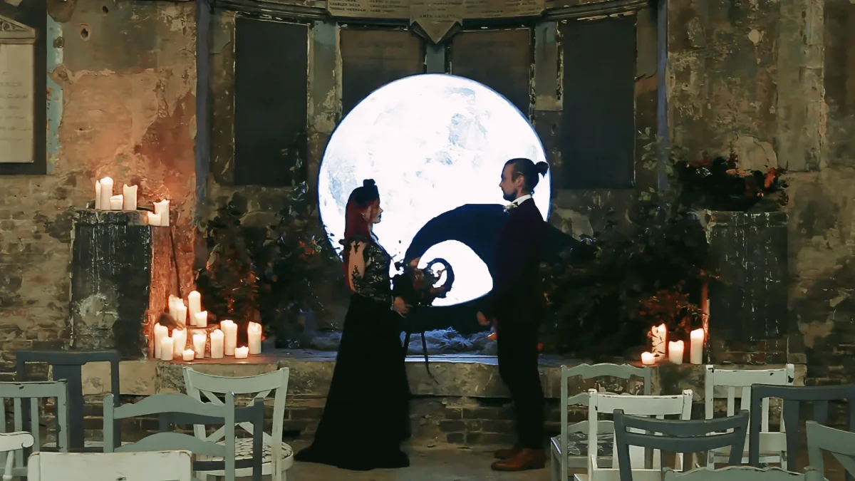 Bride and groom standing before a large moon backdrop at the Asylum Chapel in London, with candles along the aisle