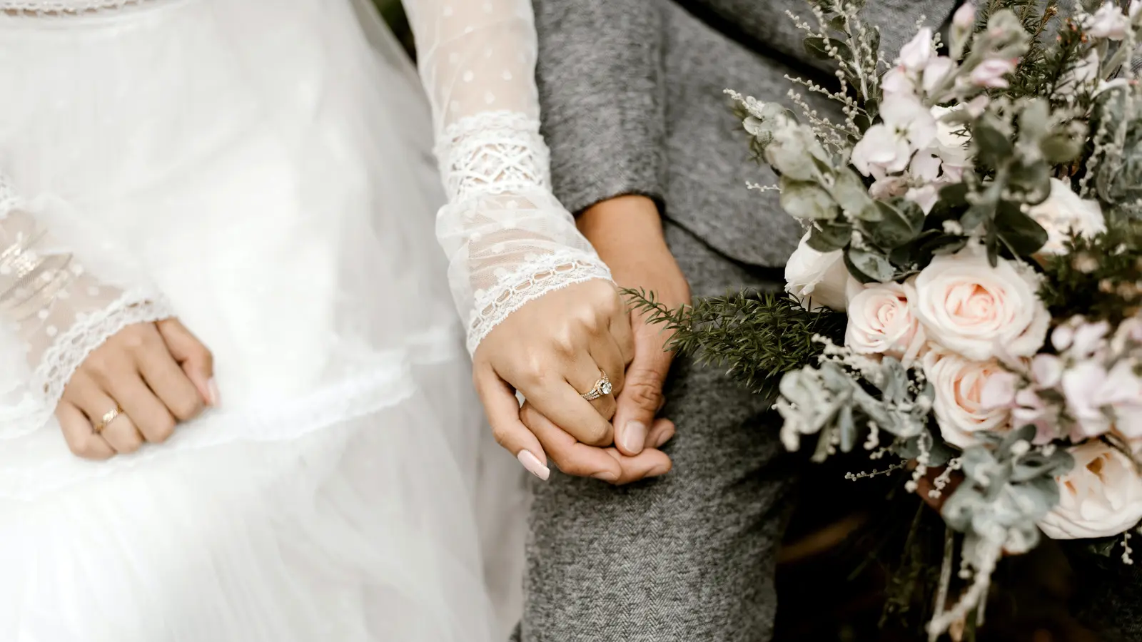 groom holds brides left hand adorned with wedding ring in his right hand while holding her bouquet