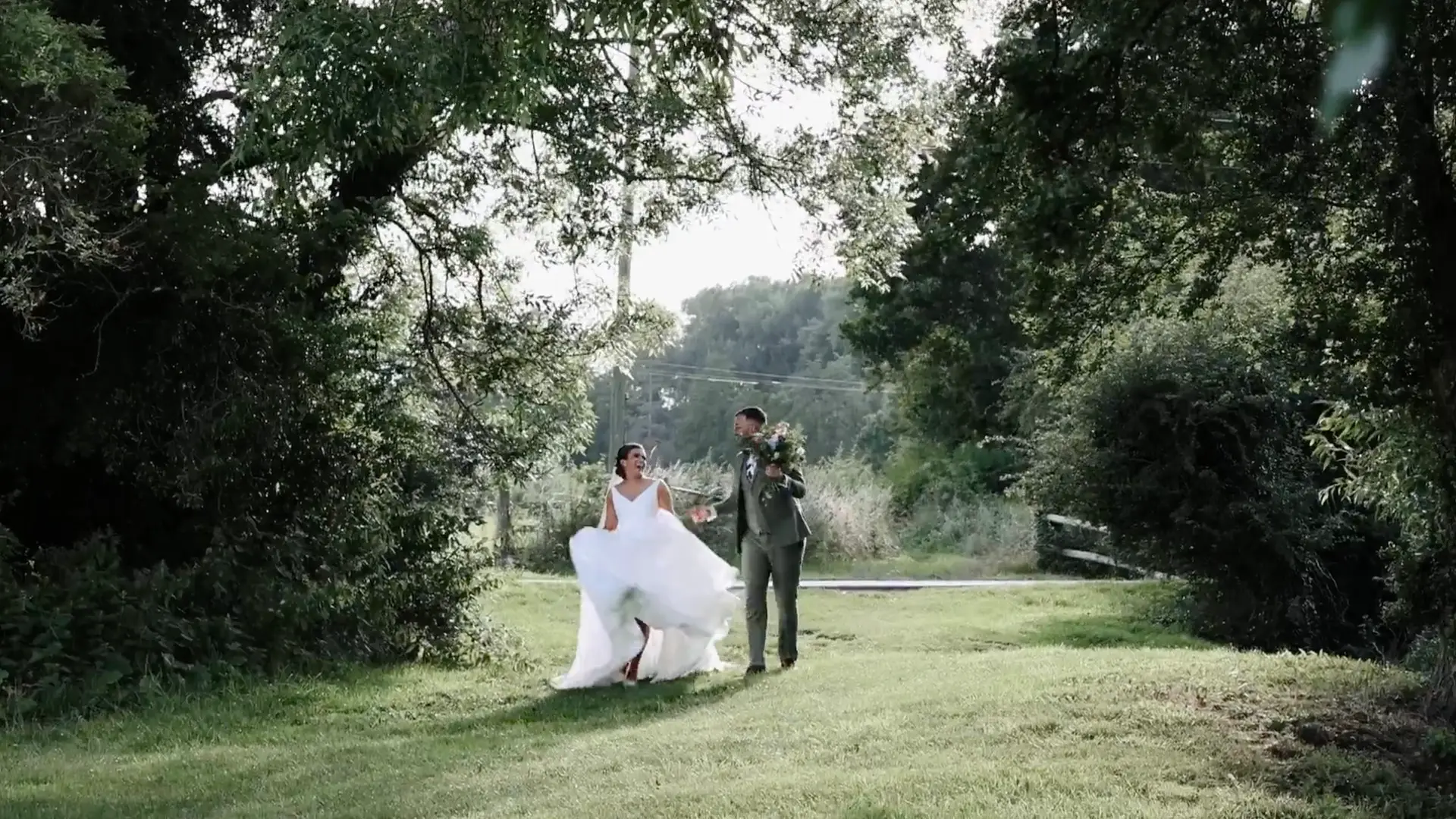 Bride and groom walk and dance through grass walkway adorned with trees either side of them, looking at each other bride sways her dress to one side and groom holds flowers
